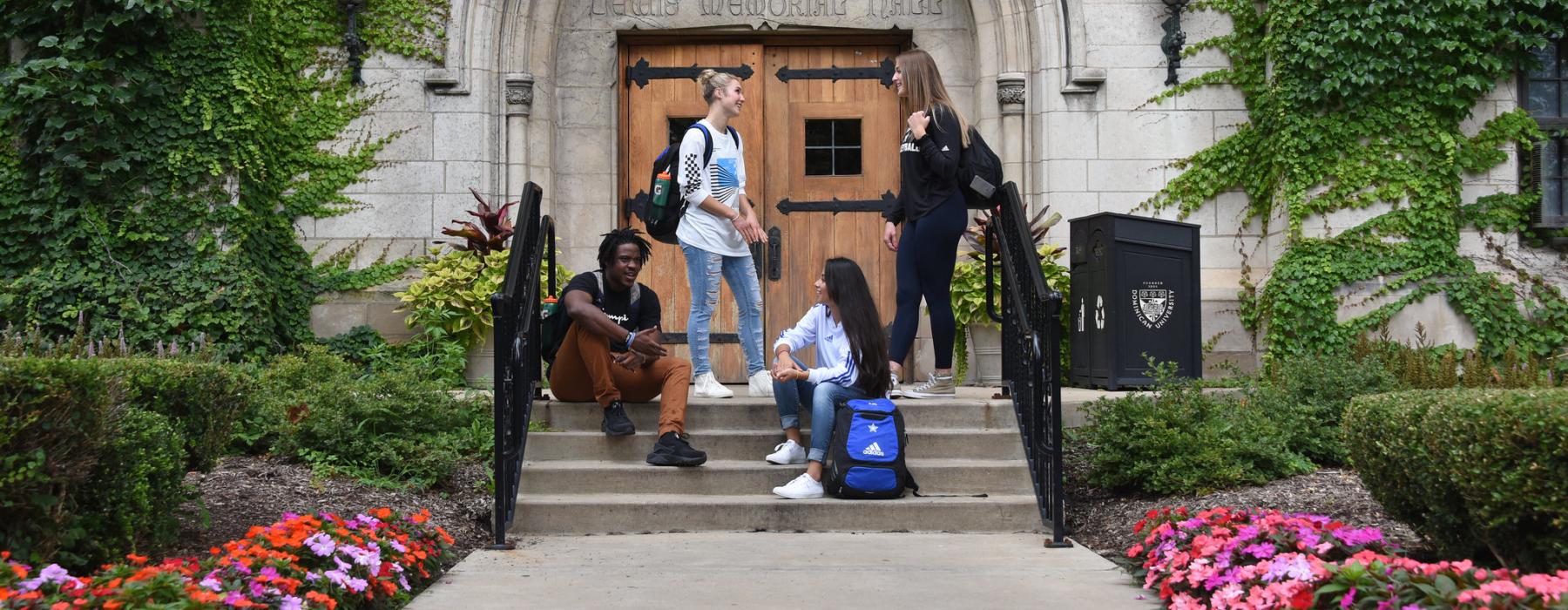 A group of four students talking in front of the Lewis Hall exterior door. Two students stand in front of the door. Two students are seated on the steps leading up to the door.